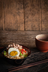Instant noodle with pork, egg and vegetables on black bowl on the wood table there are chopstick and drinking glass placed around.