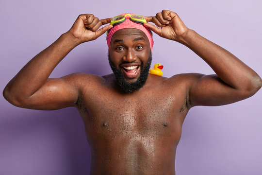 Happy dark skinned man with muscular body, adjustes goggles, smiles broadly, being wet after swimming in pool, has fun during hot summer day, has rubber duckling on strong shoulder, stands indoor