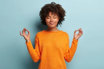 Foto op Canvas Mindful, vreedzame Afro-Amerikaanse vrouw mediteert binnen, houdt handen in mudra-gebaar, heeft ogen gesloten, probeert te ontspannen na lange uren werken, houdt vingers in yogateken, geïsoleerd op blauwe muur © Wayhome Studio
