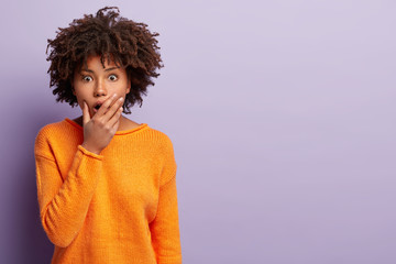 People and emotions concept. Shocked terrified woman covers widely opened mouth, hears terrible rumor, gazes with fright, wears orange jumper, models over purple background with blank space.