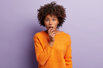 Dazed young curly female looks at camera with fright, keeps widely opened eyes and mouth, can not...