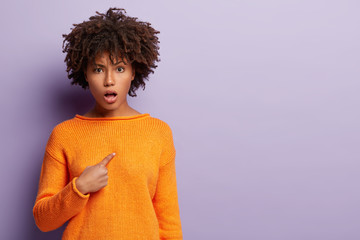 Dissatisfied woman points at herself indignantly, looks angrily at camera, discontent by unjustified claim, asks why me, wears orange jumper, stands over purple background with blank space for text