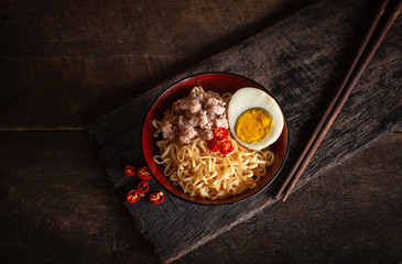 Instant noodle with pork, egg and vegetables on black bowl on the wood table there are chilli and chopstick placed around.