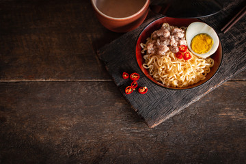 Instant noodle with pork, egg and vegetables on black bowl on the wood table there are chilli, chopstick and drinking glass placed around.