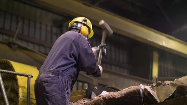 Plant worker breaks chunks of metal waste with large sledgehammer