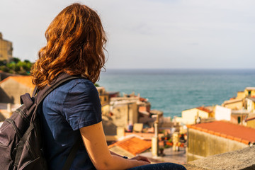 Fototapeta na wymiar Female tourist with a backpack sitting on the city walls, relaxing and enjoying the view over the colorful old town of Riomaggiore, in Cinque Terre, Italy, a famous tourist attraction.