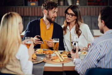 Young attractive Caucasian couple sitting in restaurant and looking at smart phone. In foreground their friends sitting adn drinking.