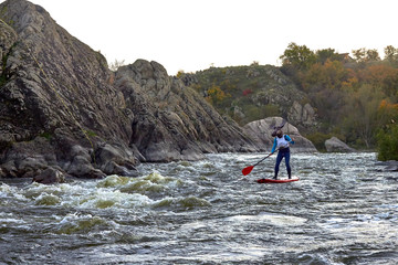 Funny man dressed as an elk (moose) is paddling on a SUP board on fast mountain whitewater river among the rapids at dusk at dusk. Costume water show. Stand up paddle boarding - active recreation.