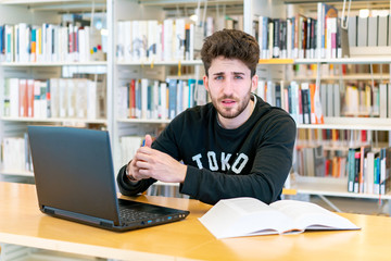 Young green-eyed student sitting in front of laptop with books at the table looking confused in the library