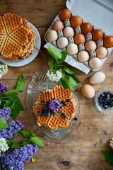 Obraz na płótnie Canvas Homemade waffles with aronia, eggs and lilac flowers on the wooden table