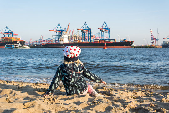 Junges Kind sitzt am Elbstrand in Hamburg mit Hafen und Schiffen im Hintergrund