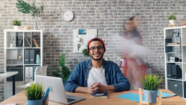 Zoom Out Time-lapse Of Happy Successful Young Man Smiling Looking At Camera In Office Room Siting At Desk When Employees Are Moving Around. Business And Youth Concept.