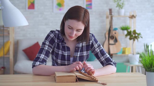 Portrait young woman sitting at a table with a rosary in her hands and reading a book