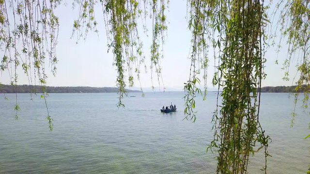 Two drone shots overlooking Lake Starnberg up to the Bavarian Alps in summer. A rowing boat is kept in view, an idyllic tree in the foreground and a great backlit reflection in the water is shown.