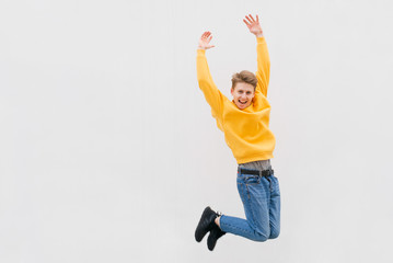 Jump of a happy young teenager in a casual dress up against a white wall, wearing jeans, yellow sweatshirt and sneakers. Joyful student jumping against the background of a white wall, street portrait