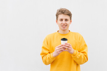 Fototapeta premium Smiling young man in casual clothing wearing a yellow sweatshirt, stands on a white background with a paper cup of coffee in his hands, looks at the camera and rejoices.