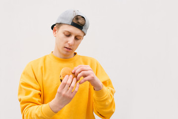 Handsome young man holding a sandwich in the background of a white wall looks like a fast food in the hands of a hungry man. Hungry teenager with a sandwich in his hands on a white background