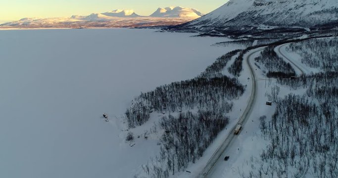 Truck on a arctic road, C4k aerial, tilt up, drone shot car driving towards mountains, boreal and polar wilderness, Tornetrask and Abisko, at sunset, on a winter evening, Lappland, Norrbotten, Sweden