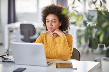 Serious mixed race businesswoman dressed casual sitting in modern office and using laptop.