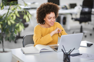 Smiling mixed race businesswoman holding eyeglasses and looking at laptop while sitting in modern...