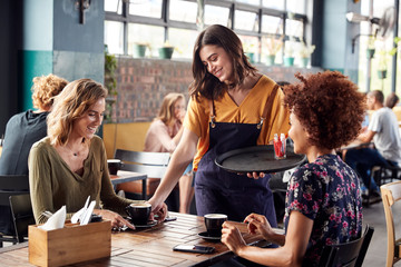 Two Female Friends Sitting At Table In Coffee Shop Being Served By Waitress - Powered by Adobe