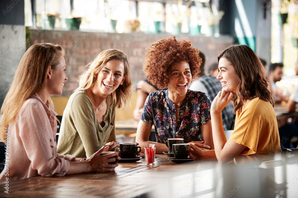 Wall mural Four Young Female Friends Meeting Sit At Table In Coffee Shop And Talk