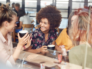 Four Young Female Friends Meeting Sit At Table In Coffee Shop And Talk
