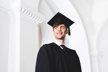 low angle view of happy young man in graduation cap smiling in university
