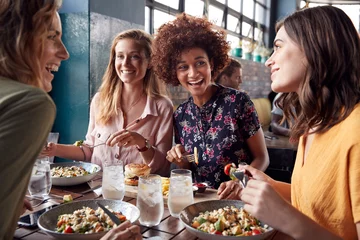 Poster Vier jonge vrouwelijke vrienden ontmoeten elkaar voor drankjes en eten en maken een toast in het restaurant © Monkey Business