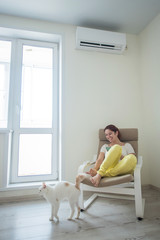 Red-haired girl in bright clothes and barefoot sits in a chair with a phone in her hands under the air conditioner. Young woman sitting in an armchair with a cat
