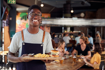 Portrait Of Waiter Serving Food To Customers In Busy Bar Restaurant - obrazy, fototapety, plakaty
