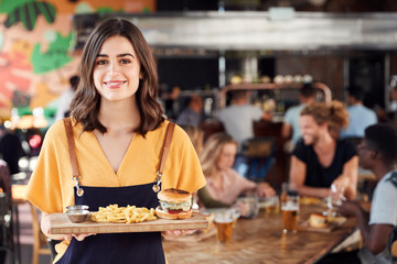 Portrait Of Waitress Serving Food To Customers In Busy Bar Restaurant - obrazy, fototapety, plakaty