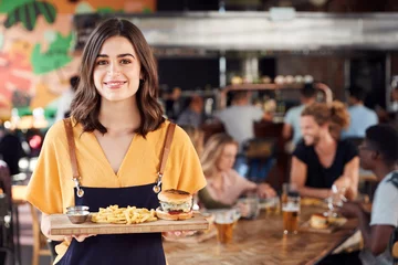 Gordijnen Portrait Of Waitress Serving Food To Customers In Busy Bar Restaurant © Monkey Business
