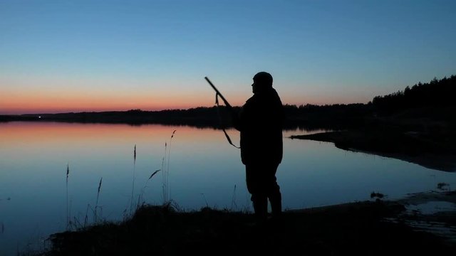 Hunter with a gun waiting for a duck at sunset by the lake. Hunting for waterfowl in the autumn-spring season.