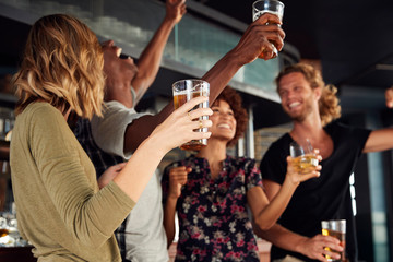 Group Of Male And Female Friends Celebrating Whilst Watching Game On Screen In Sports Bar