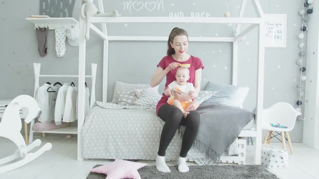 Young woman sitting on bed holding cute infant girl on knees, playful toddler daughter shoving hairbrush in mouth taking it from mom. Baby teether trying to bite comb while mother brushing her hairs.