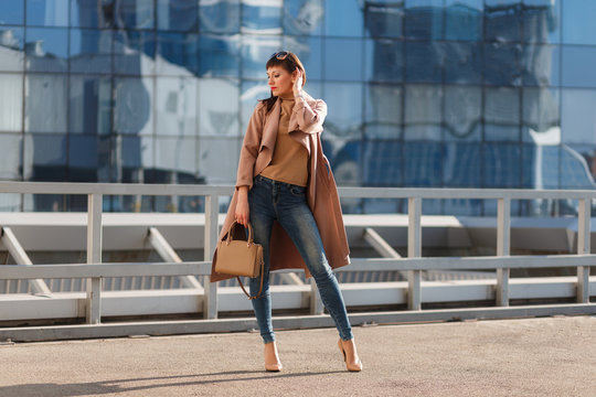 Portrait of beautiful brunette young woman in nice brown beige coat, denim jeans and sunglasses. Genuine leather bag, high heels shoes. Spring autumn fashion trendy photo of lady on urban background.