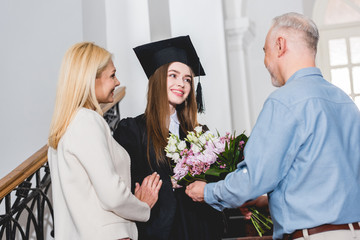 happy father giving flowers to cheerful daughter in graduation cap