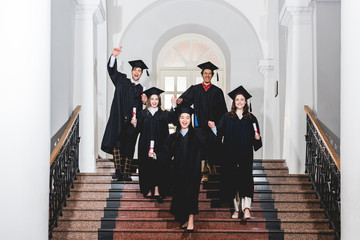 cheerful group of students in graduation gowns gesturing while holding diplomas