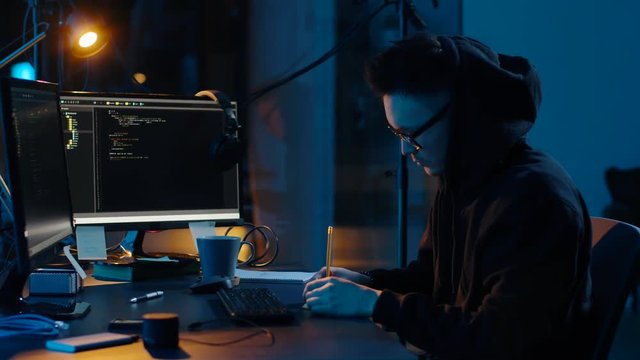 cybercrime, hacking and technology concept - asian male hacker in hoodie using computer virus program for cyber attack and taking notes in dark room at night
