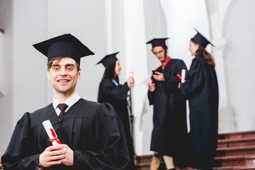 selective focus of happy man in graduation gown looking at camera and holding diploma near students