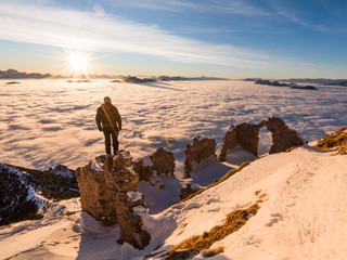 Man standing on a ruin on the top of a mountain in winter with snow and a valley full of fog at sunset with the sun
