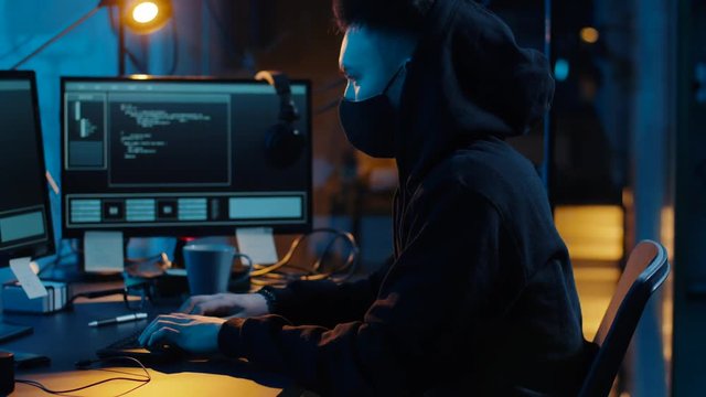 cybercrime, hacking and technology concept - asian male hacker in mask writing code or using computer virus program for cyber attack in dark room at night