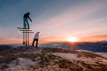 Couple on the top of a mountain peak at with the men standing on the top and giving the hand to his girlfriend at sunset / sunrise in winter with snow