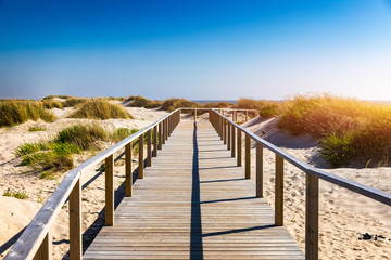 Fototapeta na wymiar Wooden path at Costa Nova d'Aveiro, Portugal, over sand dunes with ocean view, summer evening. Wooden footbridge of Costa Nova beach in a sunny day. Aveiro, Portugal.