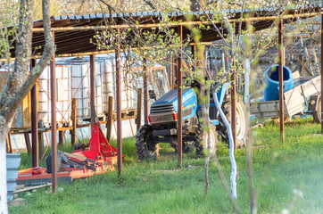 Fototapeta na wymiar Tractor on a small home farm for vegetables and fruits. Green grass, blooming plum trees. Blue tractor with rammers and milling machines for land processing. Rural home farm.