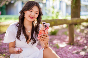 A young beautiful Asian woman in a white dress walks in a flowered park. Young beautiful asian woman making selfie on phone. Sakura. Blooming trees.