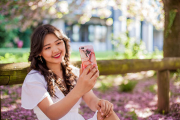 A young beautiful Asian woman in a white dress walks in a flowered park. Young beautiful asian woman making selfie on phone. Sakura. Blooming trees.