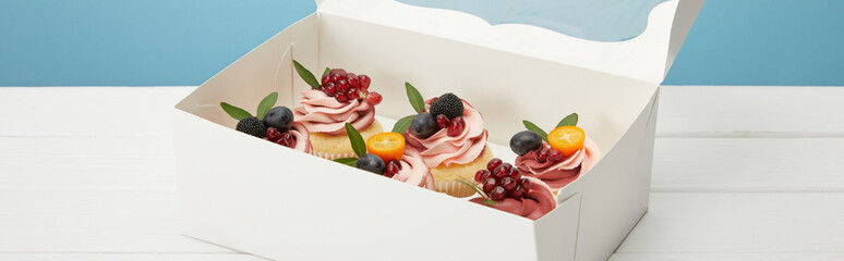 panoramic shot of cupcakes with berries and fruits in box on white surface isolated on blue