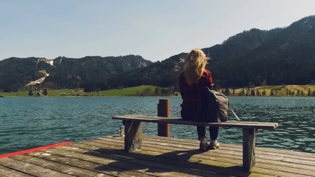 Young girl with backpack is sitting on bench at the pier on mountain lake against the background of mountains. Summer, sunny day, girl is resting on the pier on mountain lake.